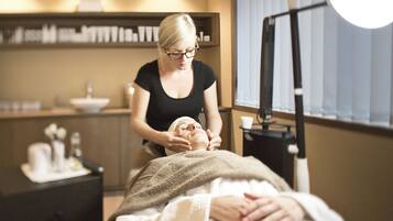 A person receiving a facial treatment in a room with a bed, a nightstand, and a shelf with toiletries.