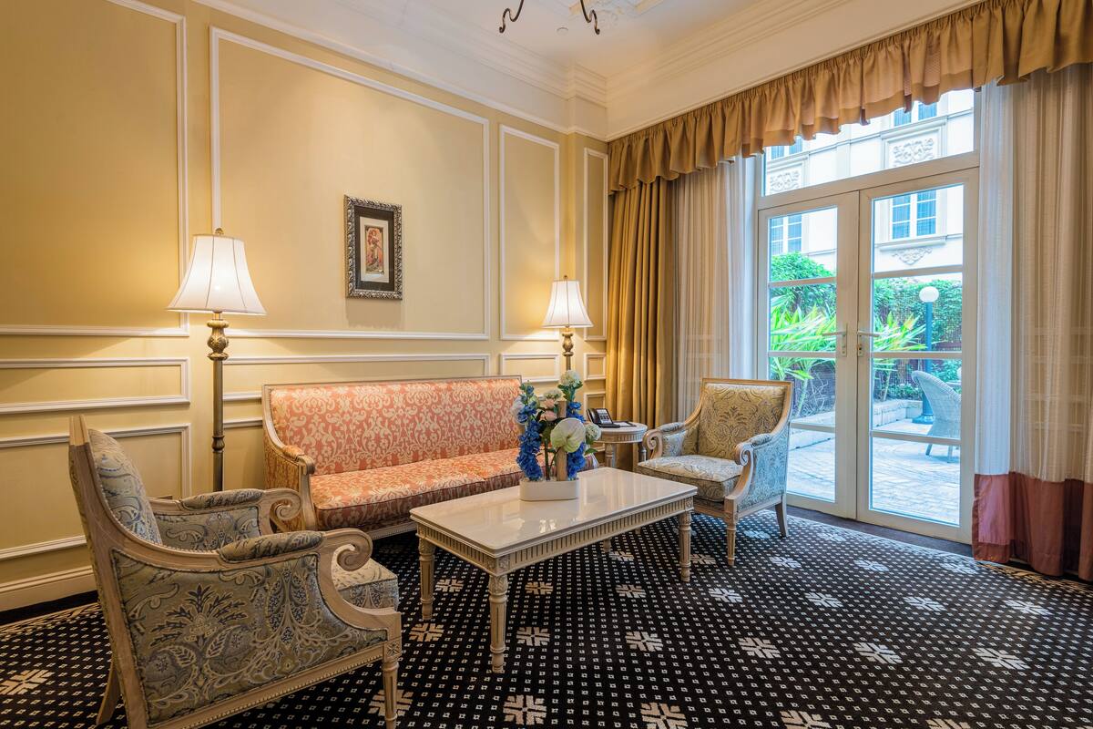 Ein Zimmer mit einem gemusterten Boden, einem Sofa, zwei Sesseln, einem Tisch mit Blumen und Blick ins Freie durch eine Glastür.