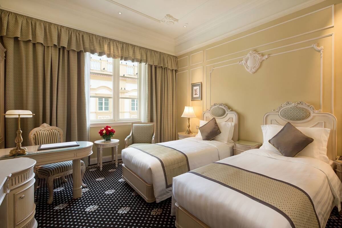 Ein Hotelzimmer mit zwei Betten, einem Schreibtisch, einem Stuhl, einer Lampe und einem Fenster mit Vorhängen.