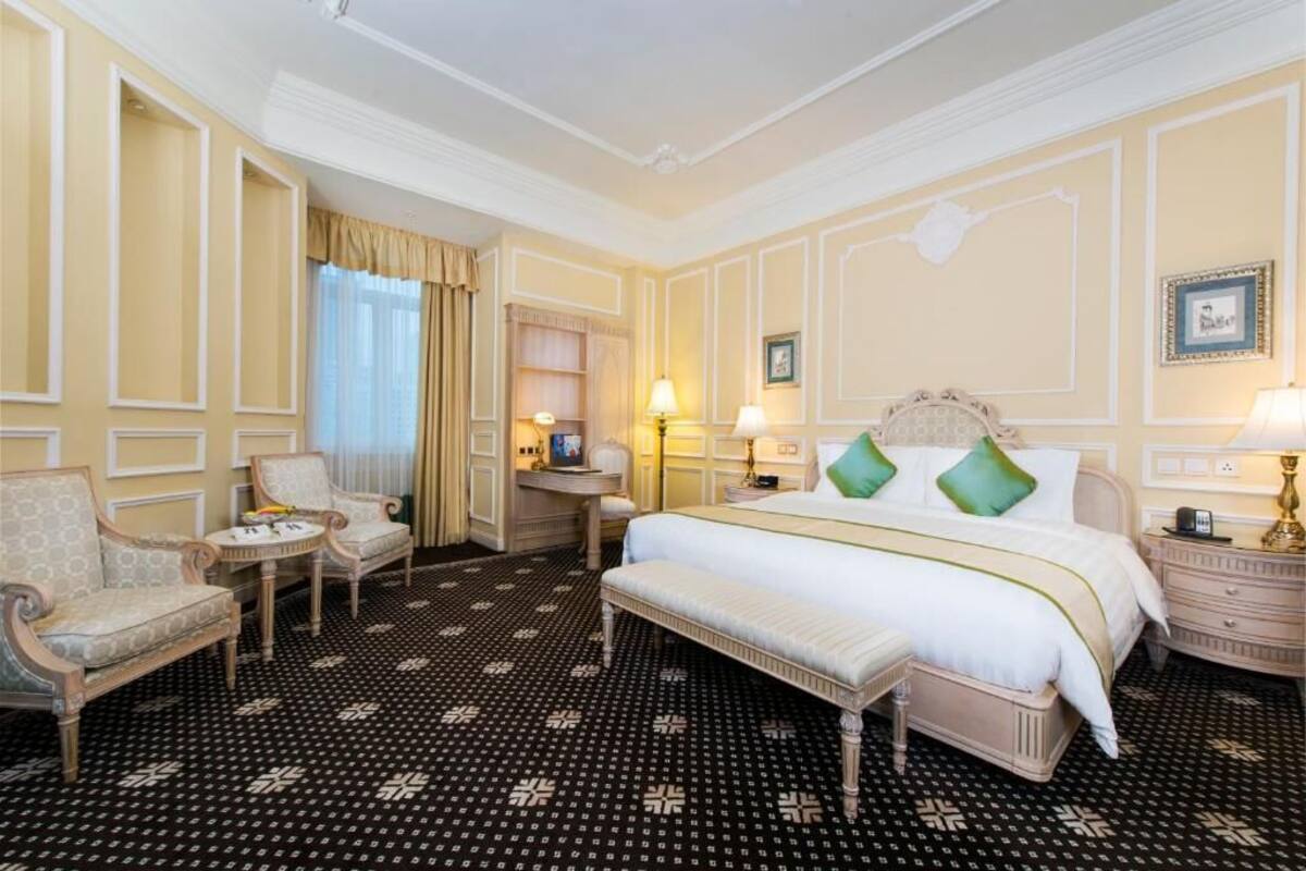 Ein Hotelzimmer mit einem großen Bett, zwei Nachttischen, einer Bank, einem Sessel, einem kleinen Tisch und einem Fenster mit Vorhängen.