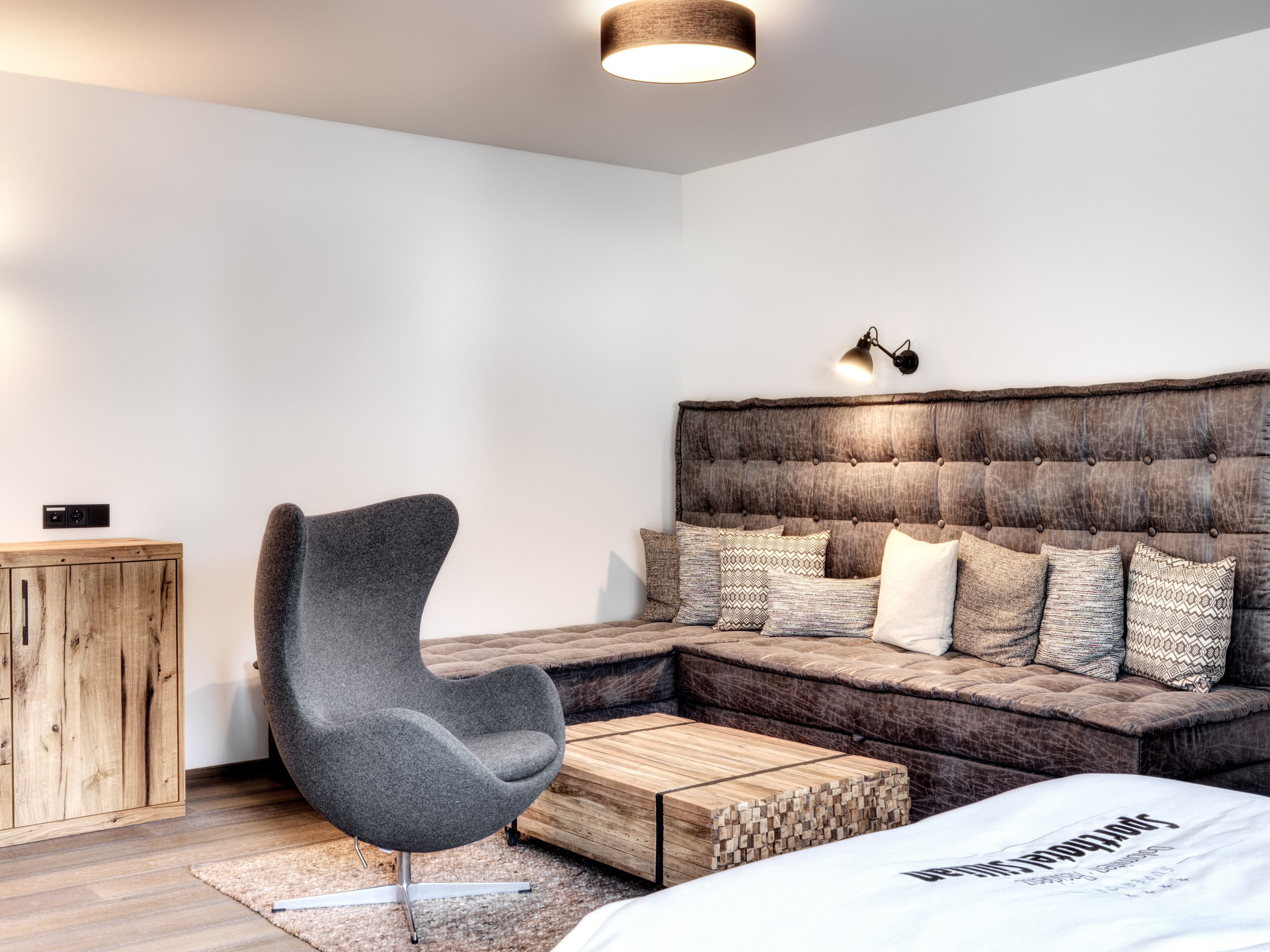 A modern bedroom with a sectional sofa, a grey armchair, a wooden coffee table, and a bed with a white comforter.
