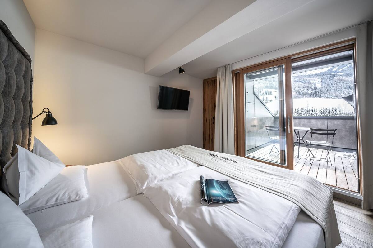 Une chambre avec un lit, un téléviseur fixé au mur et une vue sur une terrasse en bois à l’extérieur.