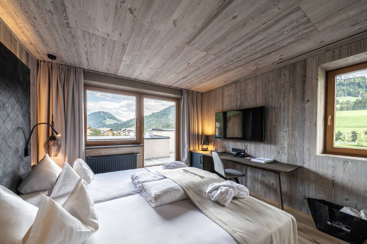 Une chambre d’hôtel avec un plafond en bois, un grand lit avec du linge de maison blanc, un bureau avec un ordinateur, une télévision et une vue sur les montagnes par la fenêtre.