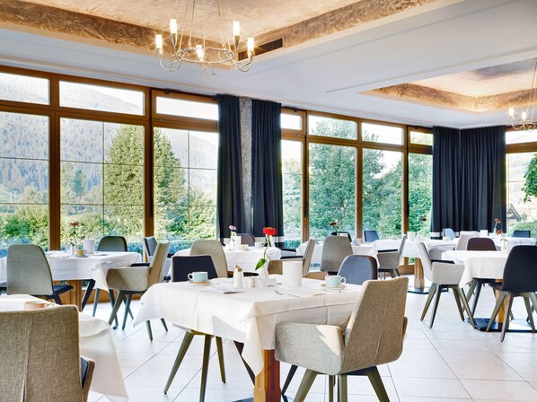 Un espace repas comprenant plusieurs tables et chaises, de grandes fenêtres offrant une vue sur les montagnes et un lustre.