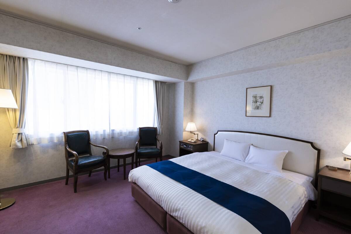 A hotel room with a large bed, two armchairs, a small table, a nightstand with a lamp, and a window with curtains.