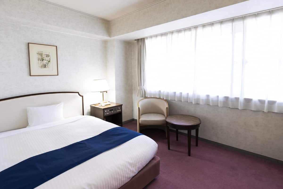 A hotel room with a large bed, a small table, a chair, a nightstand, a lamp, and a window with curtains.