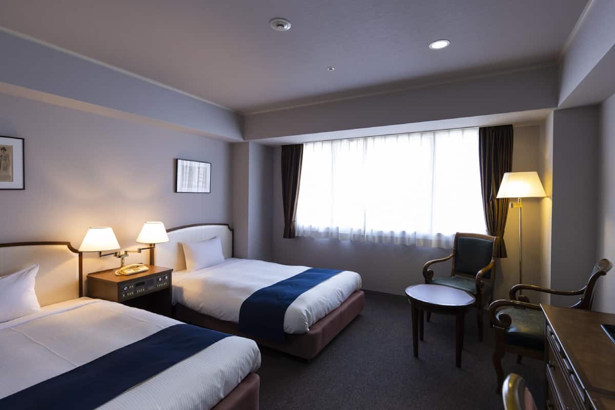 A hotel room with two beds, a desk, a chair, and a window with curtains.
