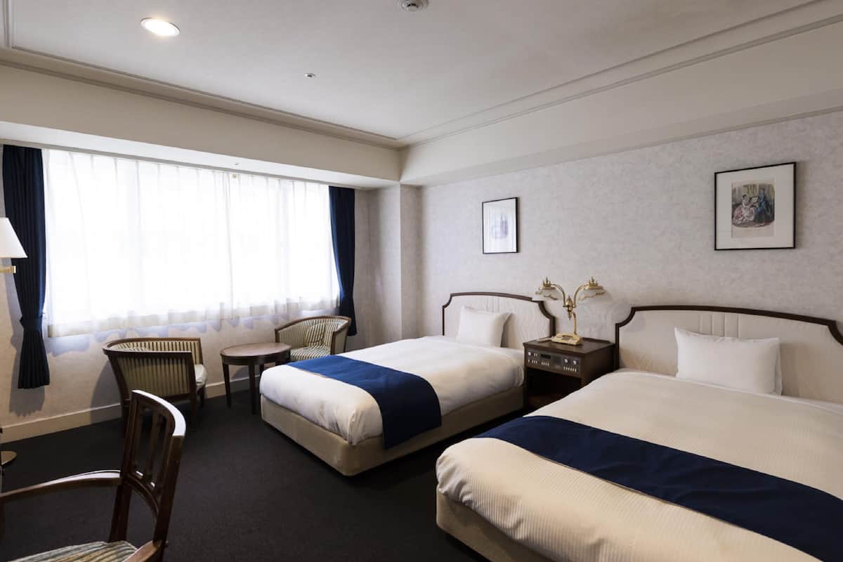 A hotel room with two beds, a small table, a chair, and a window with curtains.