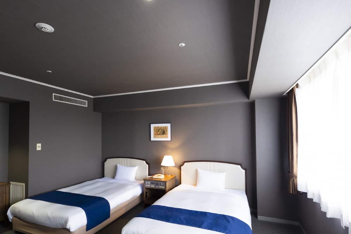 A hotel room with two beds, a bedside table with a lamp, a picture on the wall, and a window with curtains.