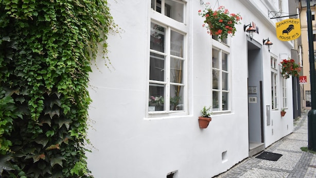 Um edifício branco com hera à esquerda, janelas adornadas com vasos de flores pendentes e uma placa amarela indicando um prédio de apartamentos.