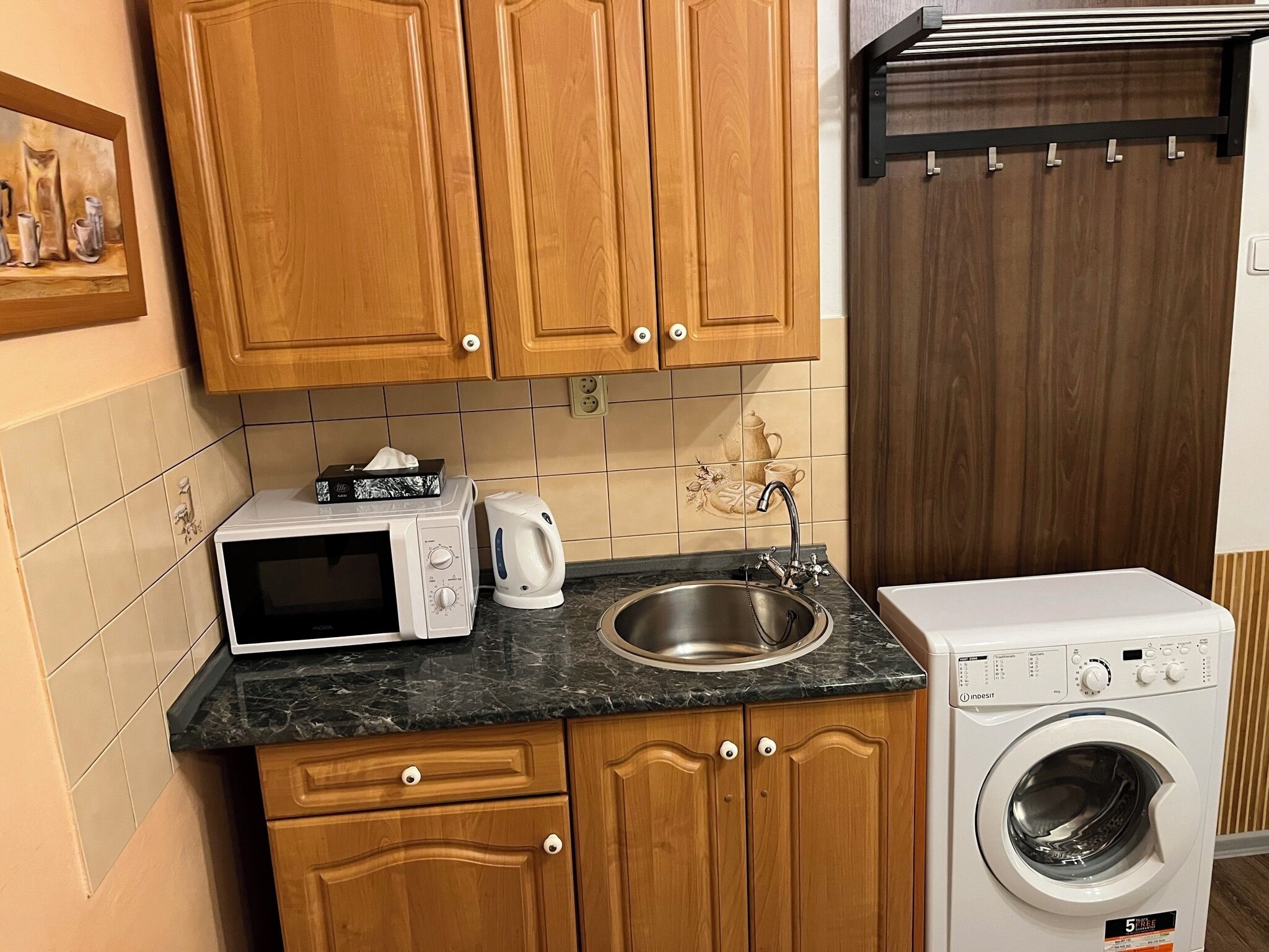A kitchen with wooden cabinets, a microwave, a kettle, a sink, and a washing machine.