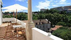Una terraza con piso a cuadros, mesas y sillas blancas, un paraguas blanco y vistas a edificios y árboles.
