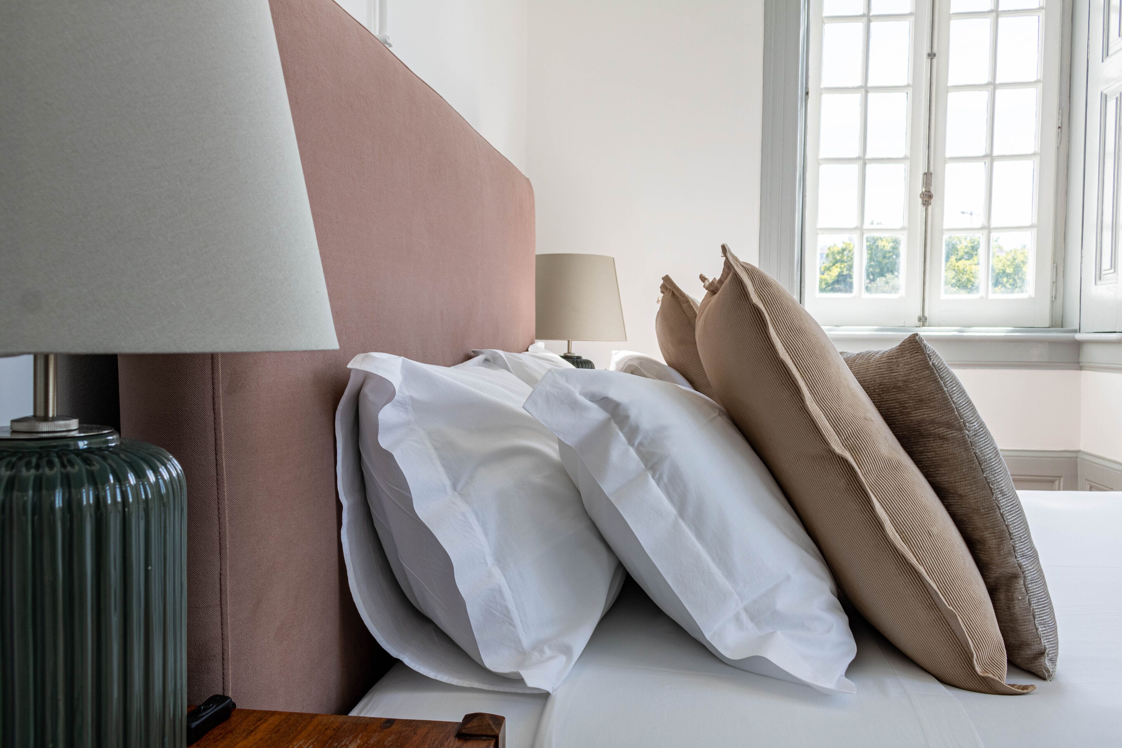 A neatly made bed with white and beige pillows, a green lamp, and a window with a view of trees.