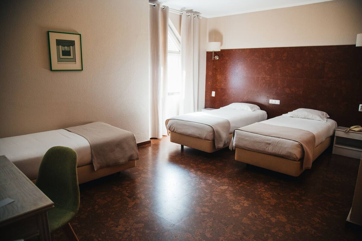 Une chambre d’hôtel avec deux lits simples, un bureau, une chaise et une fenêtre avec des rideaux.
