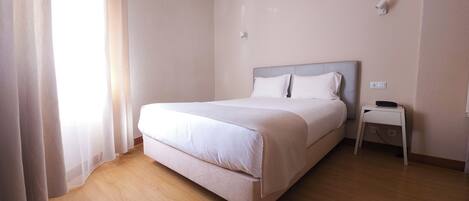 Cama bien hecha en una habitación de hotel, con suelo de madera, una mesita de noche con teléfono y una lámpara de pared.