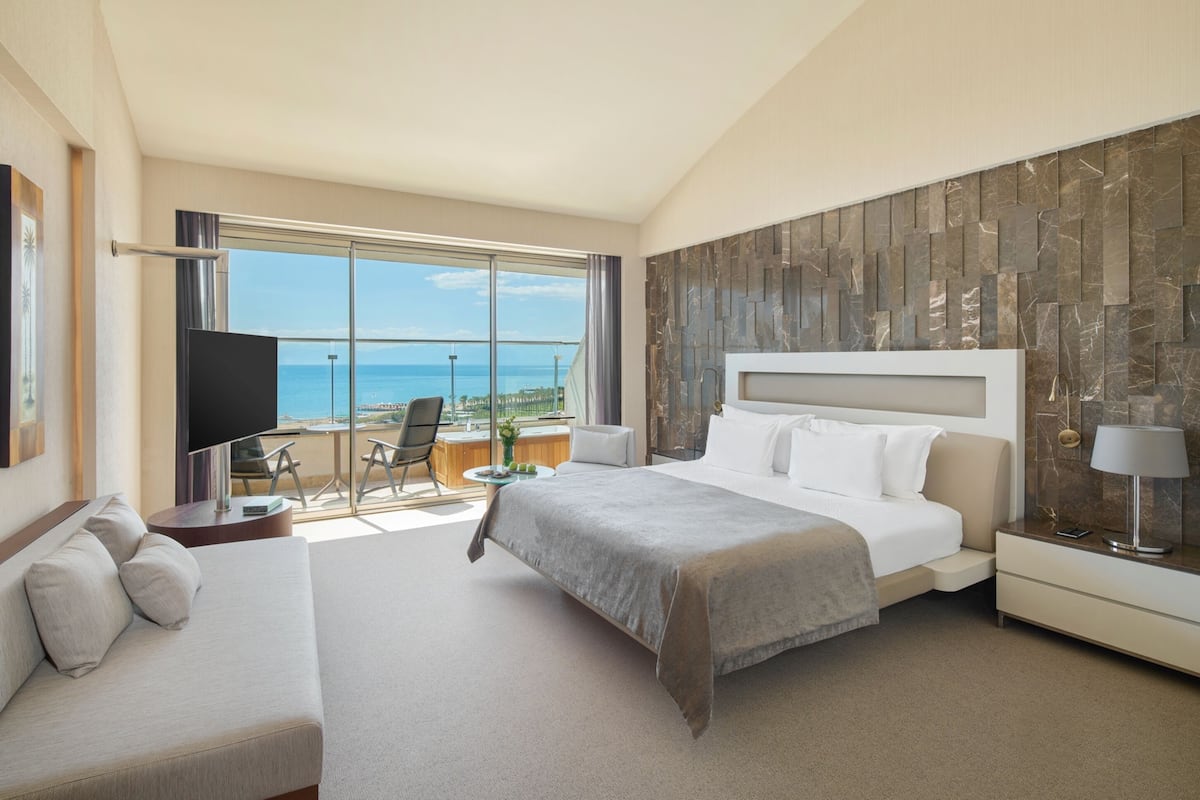 A modern hotel room with a large bed, a sofa, a TV, and a balcony with a view of the sea.