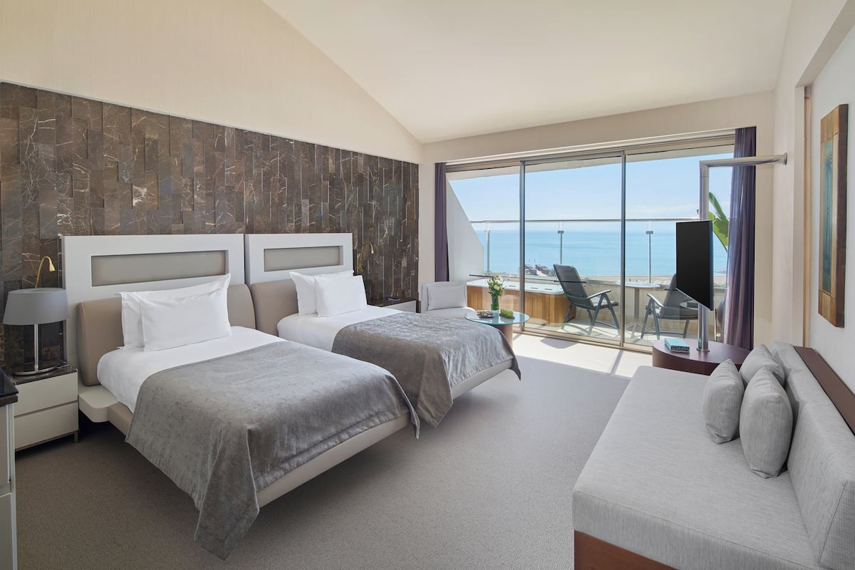 A modern hotel room with two beds, a sofa, a balcony with a view of the sea, and a flat-screen TV.