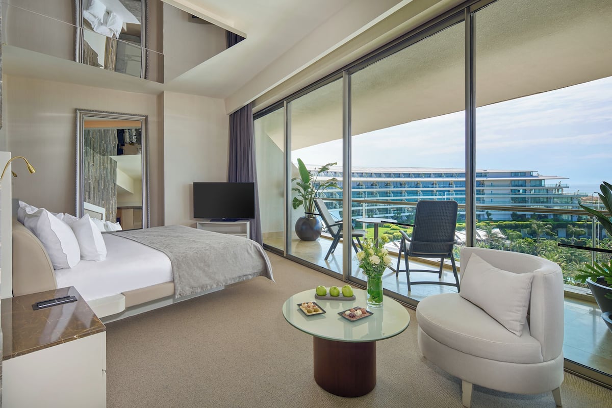 A modern hotel room with a bed, a TV, a glass table with snacks, a sofa, and a balcony with a view of a building and greenery.