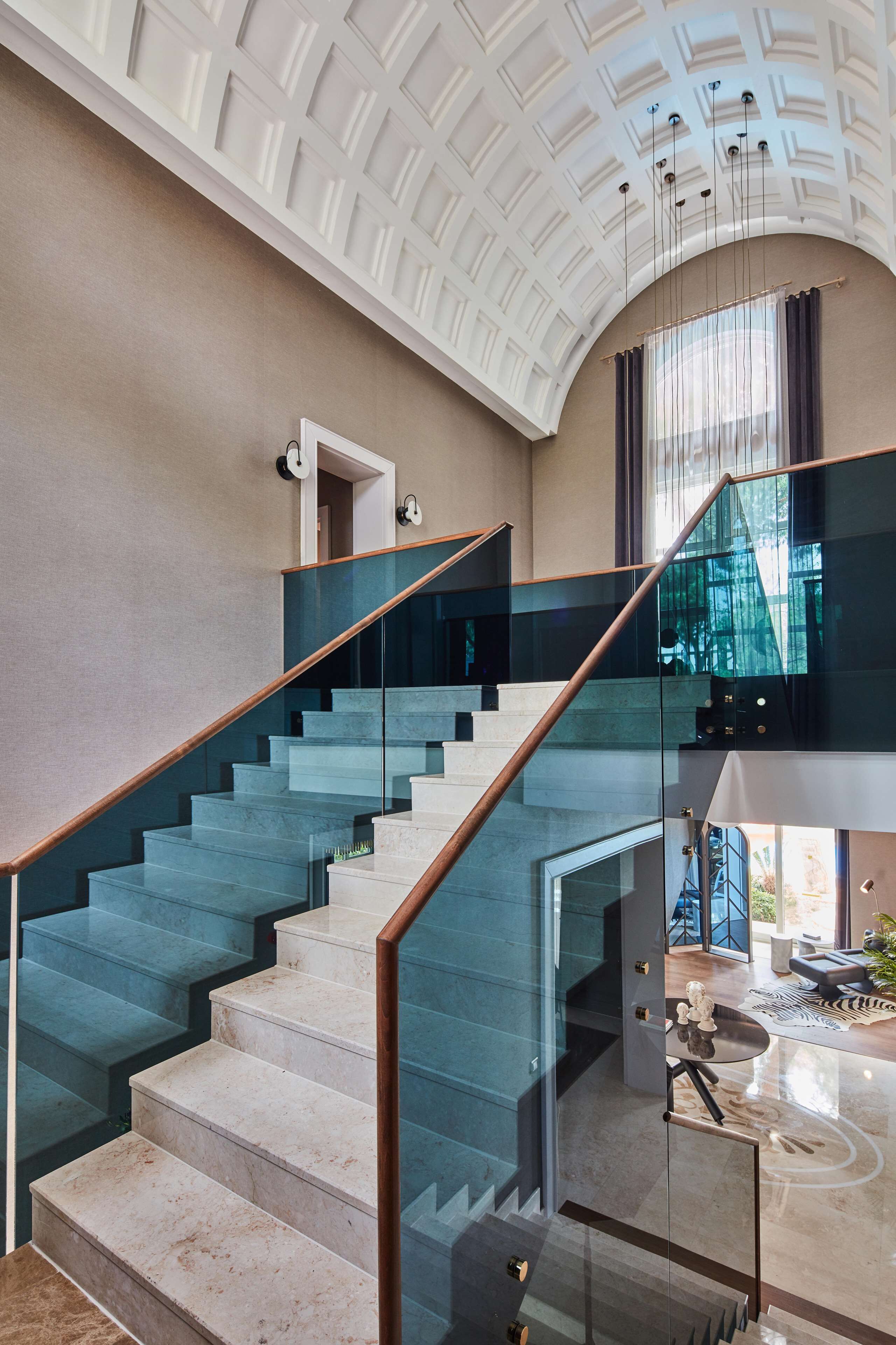 Una escalera moderna con barandillas de vidrio y barandilla de madera que conduce a un segundo piso con vistas al exterior.