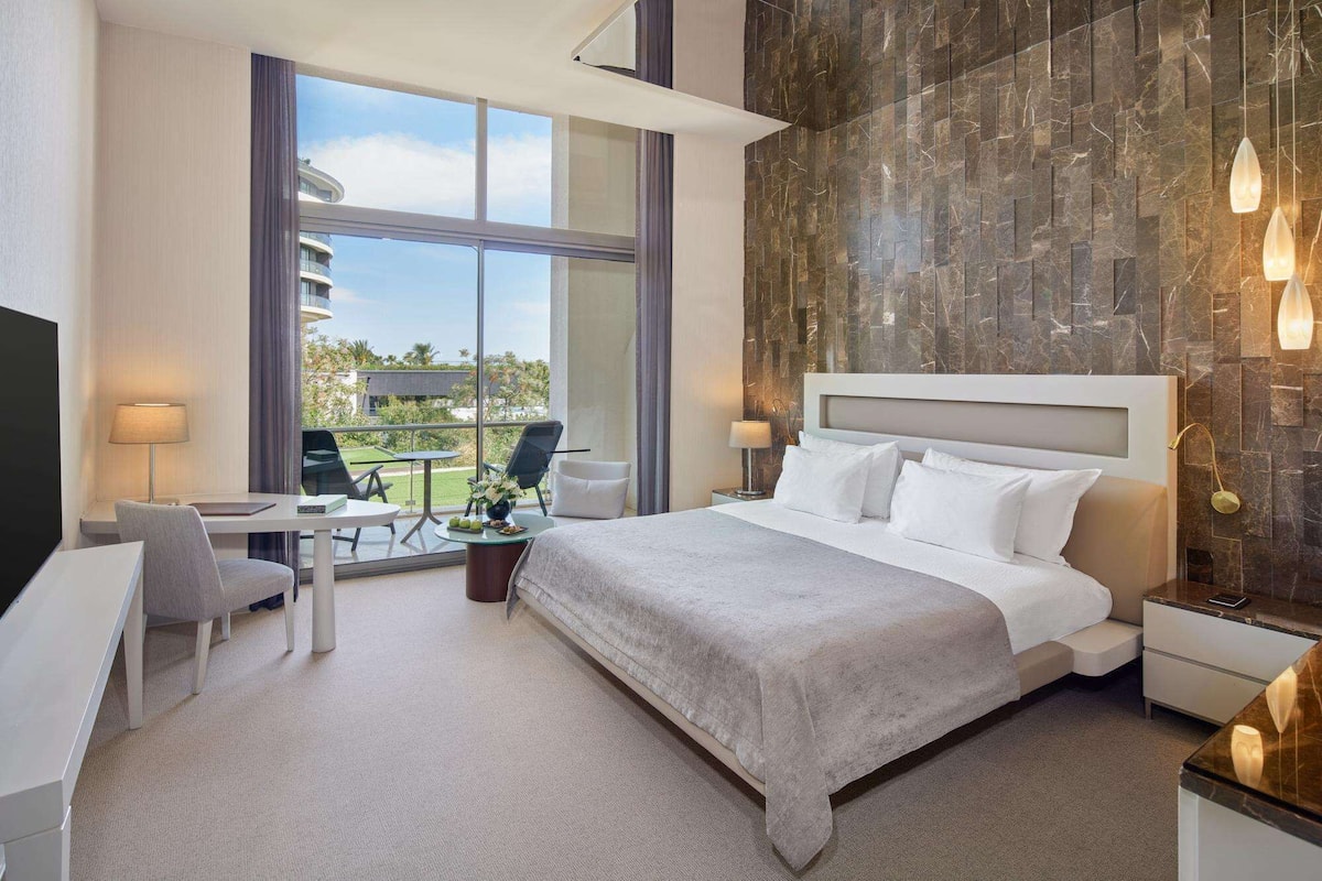 A modern hotel room with a large bed, a desk, and a chair. There is a view of the outdoors through large windows.