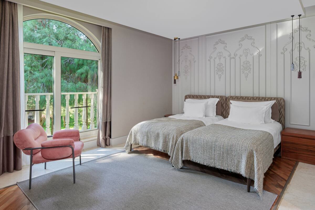 A bedroom with two beds, a large window with curtains, a pink chair, and a wooden floor.