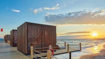 Eine Holzstege führt zu einer Reihe moderner Strandhütten aus Holz, mit einer Flagge, einem Rettungsring und einem hölzernen Steg über dem Wasser.