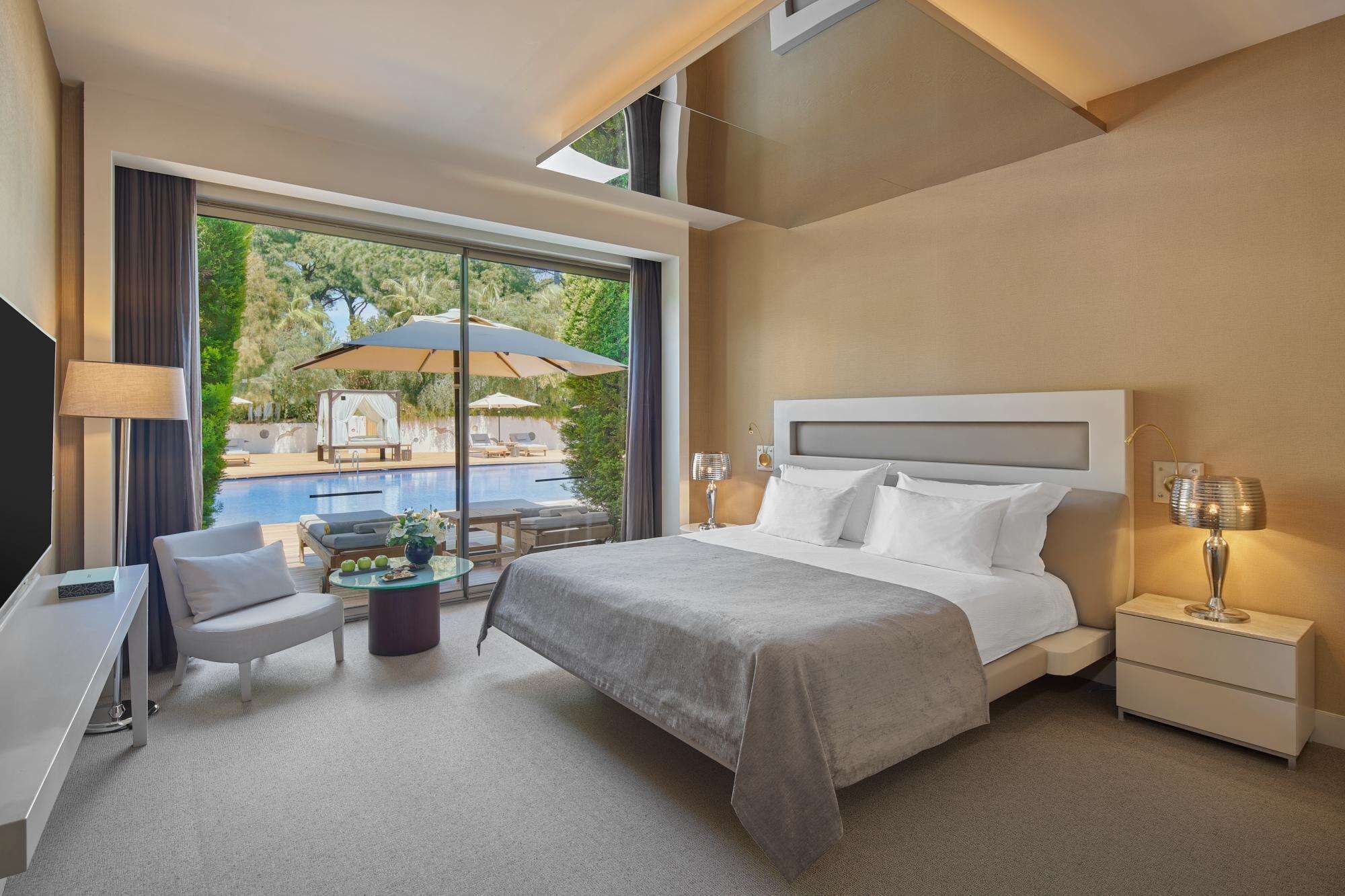 A modern hotel room with a large bed, a desk, a chair, a small table, and a view of an outdoor pool and greenery.