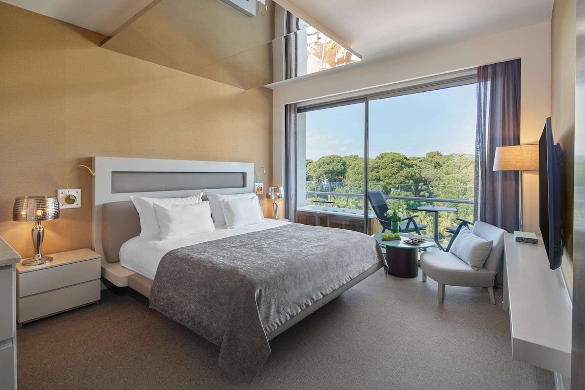 A hotel room with a large bed, a TV, a balcony with a view of trees, and a chair.