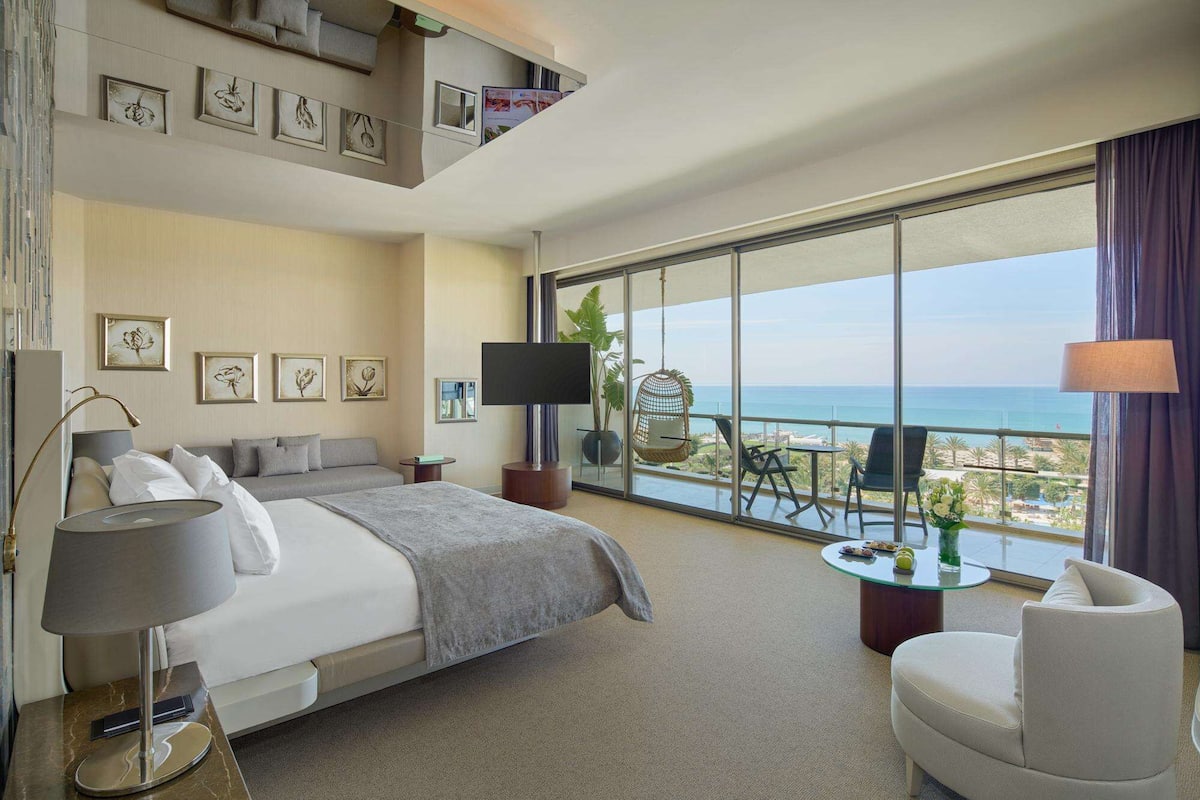 A hotel room with a large bed, a sofa, a TV, a balcony with a view of the sea, and a chair.