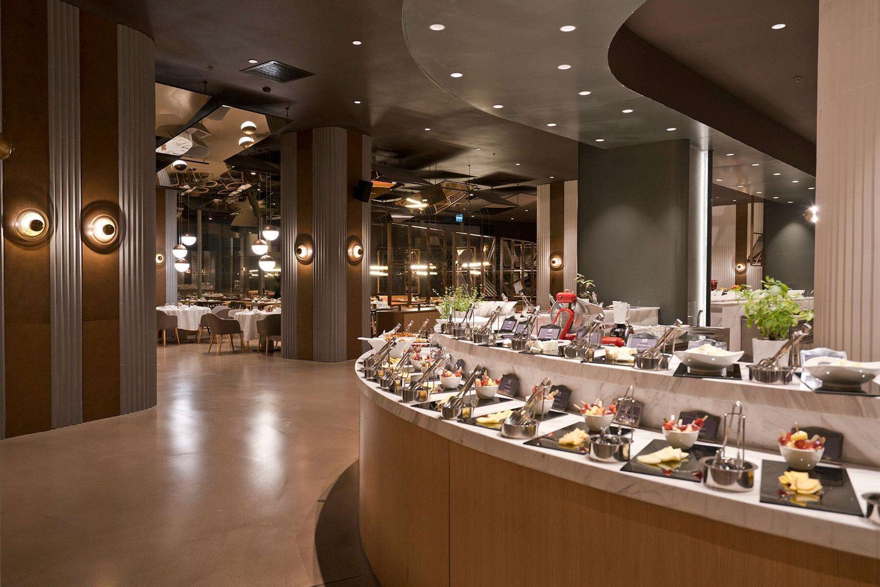 Een modern restaurant met een buffetopstelling, met een gebogen toonbank vol diverse gerechten en bestek.