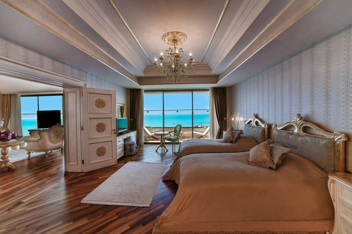 A luxurious bedroom with a large bed, a chandelier, a desk with a TV, and a view of the ocean.
