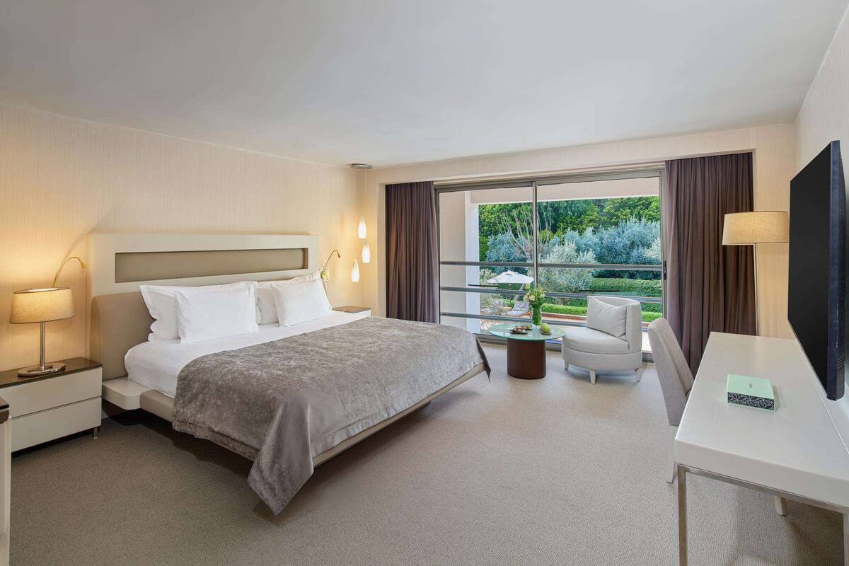 A hotel room with a bed, bedside tables, a desk, a chair, a TV, and a window with a view of greenery.