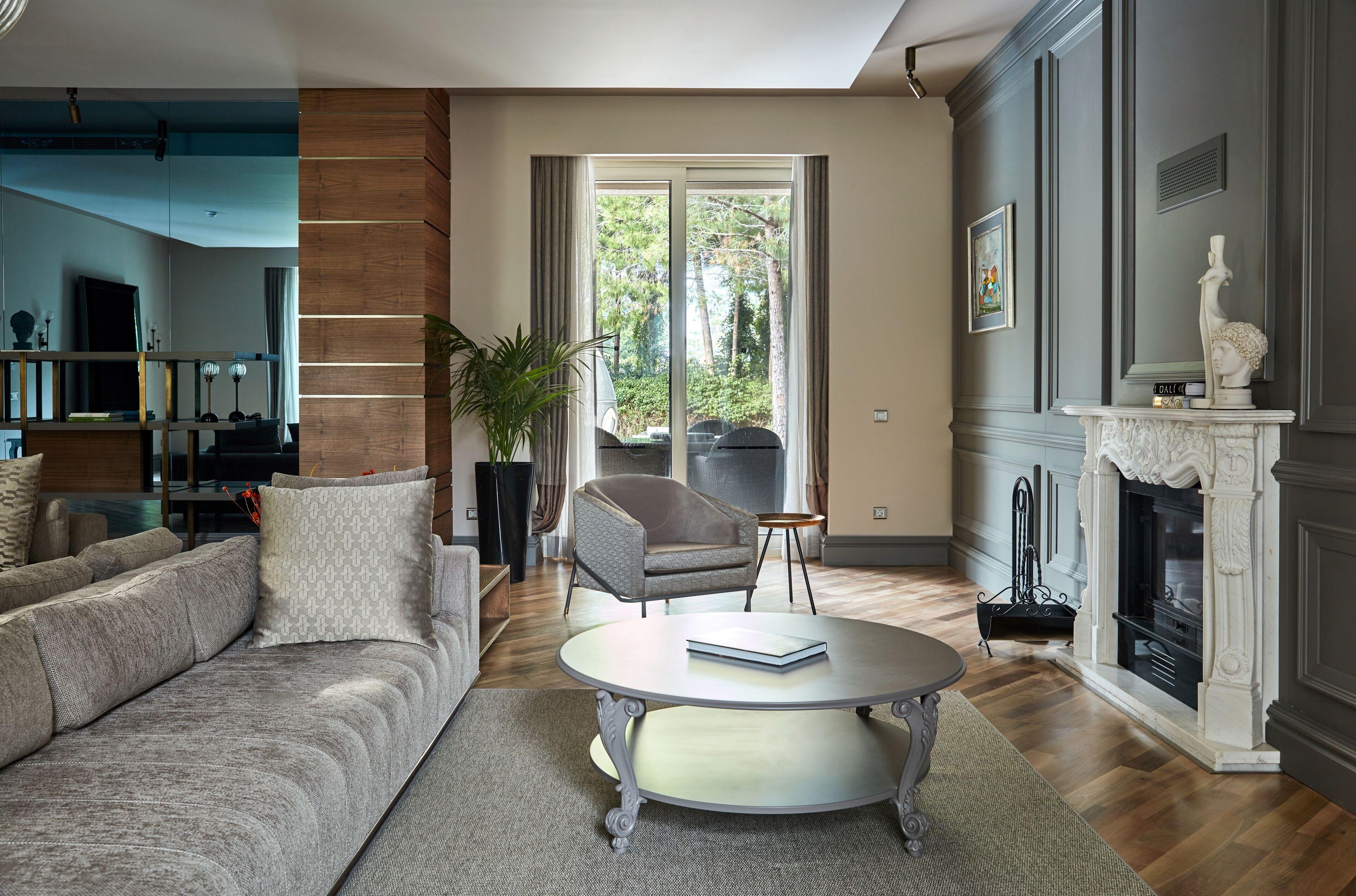 Un salon avec un canapé gris, une table basse ronde, une cheminée et une grande fenêtre donnant sur la forêt.