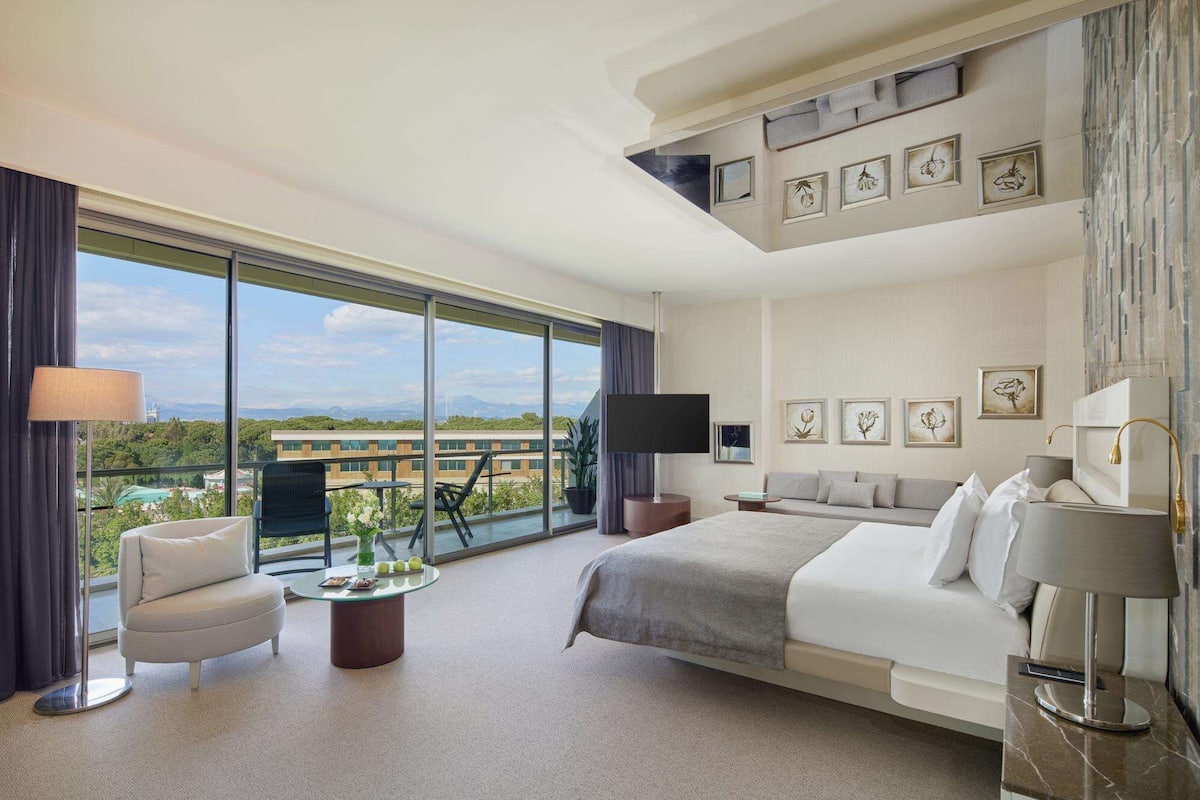 A hotel room with a large bed, a sofa, a round table with a lamp, and a TV. There are framed pictures on the wall and a view of the city through large windows.