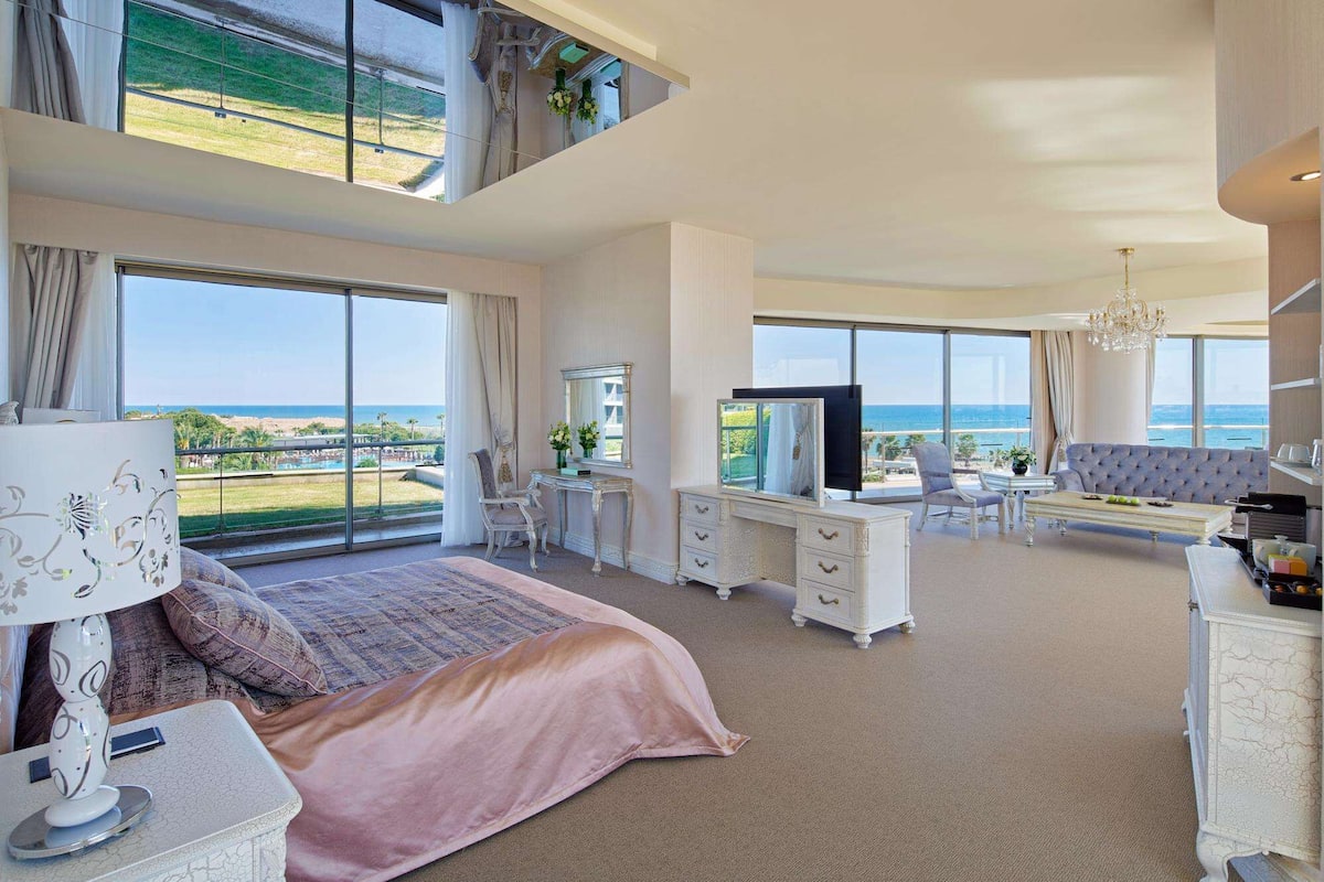 A spacious bedroom with a large bed, a desk, a television, and a view of the ocean.