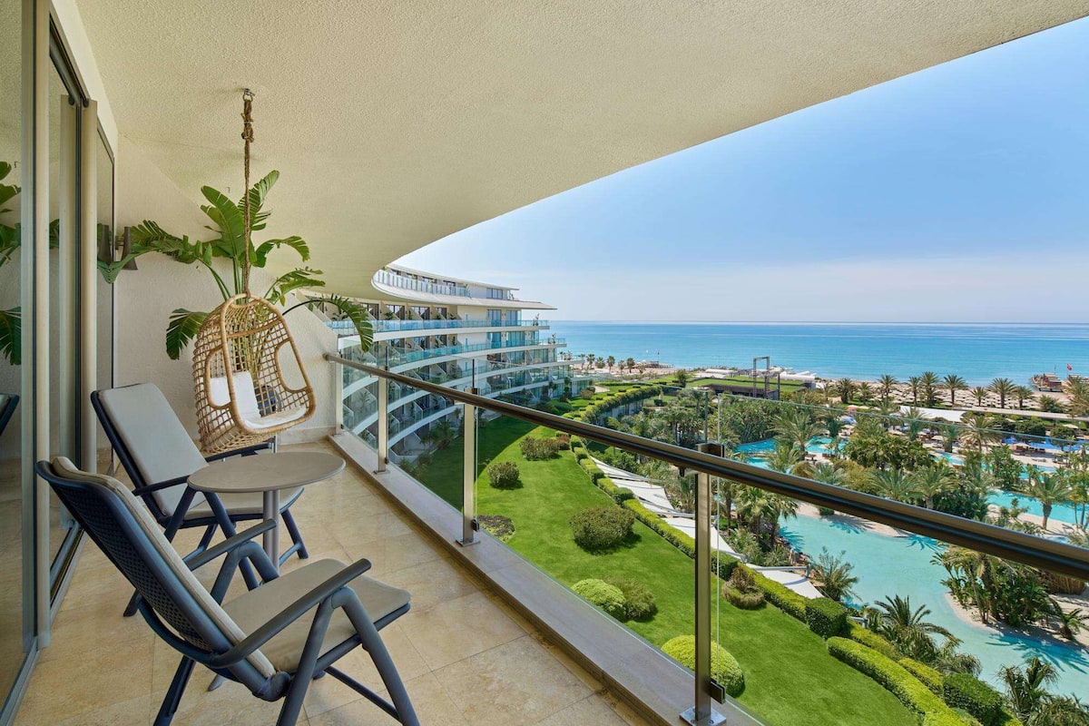 A balcony with two lounge chairs, a hanging chair, and a view of a resort with a pool and beach.