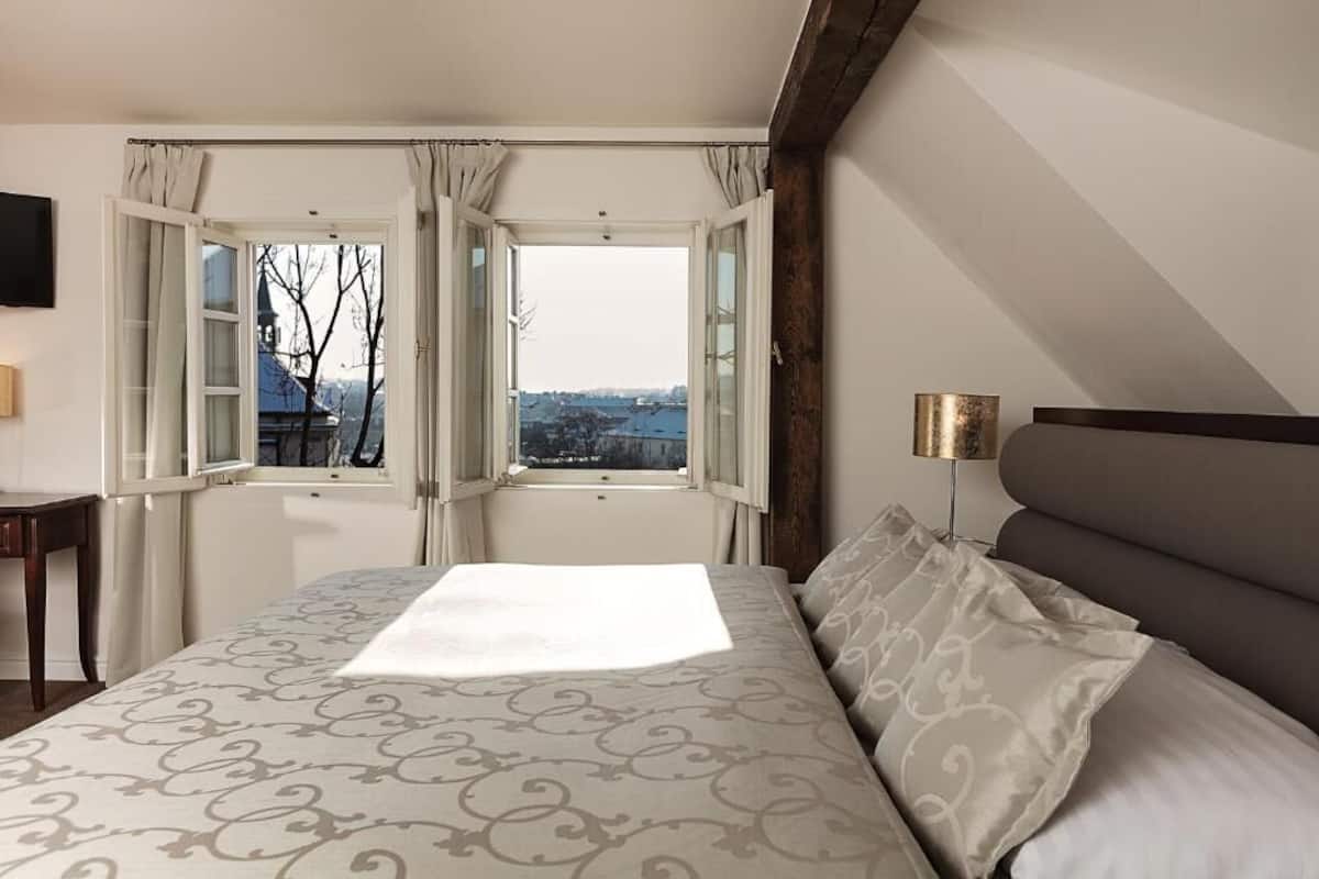 A bedroom with a large bed, a view through an open window, and a bedside table.