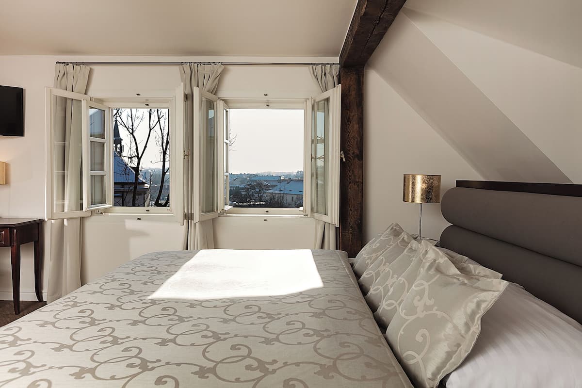 A bedroom with a large bed, a nightstand, a lamp, and a view of the city through open windows.