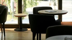 Interior de una cafetería con un ventanal grande, dos mesas redondas y sillas negras.