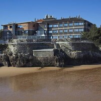 Un hotel de varios pisos con fachada de piedra, un balcón y una playa arenosa al frente.