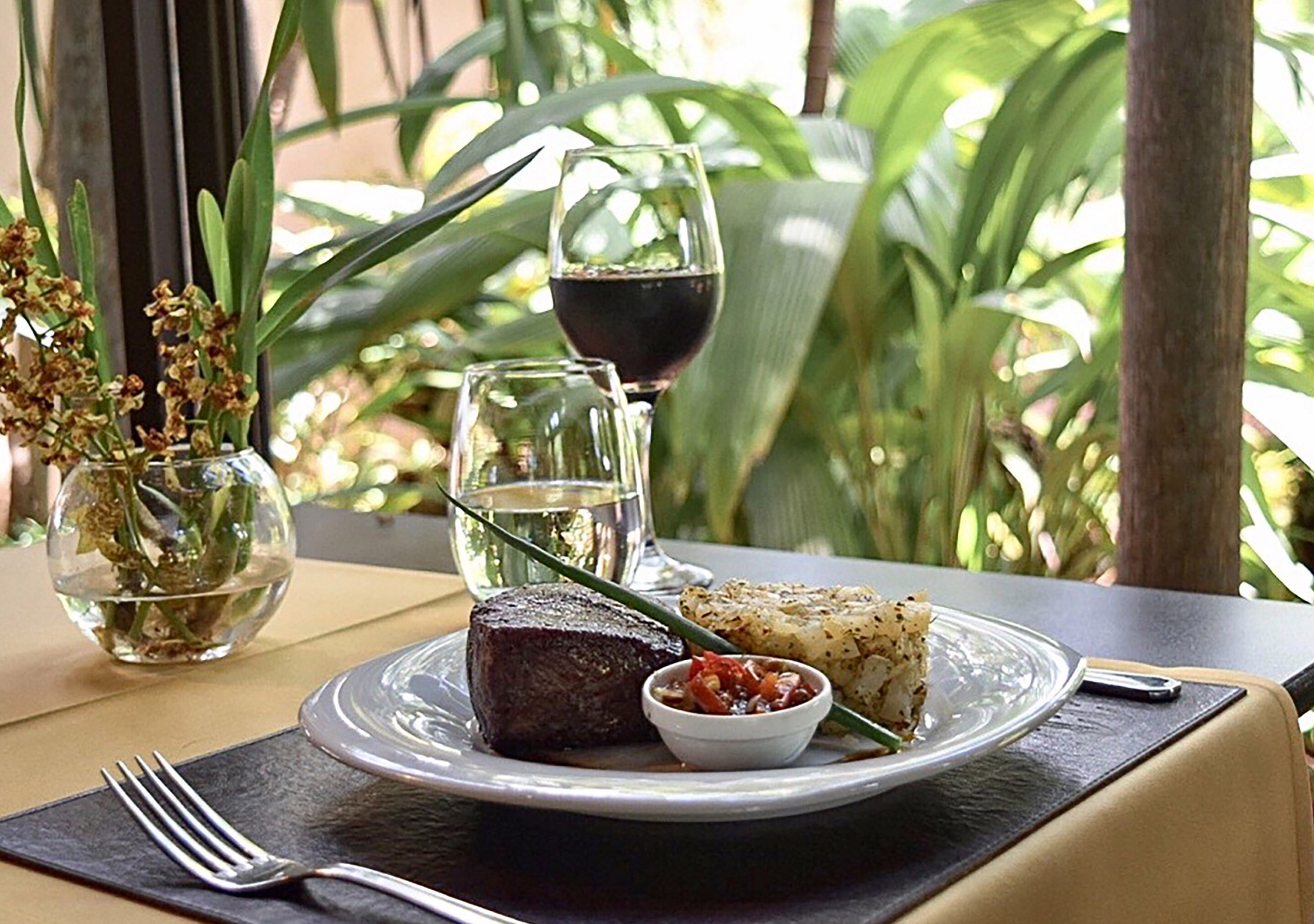 A table set with a steak, a glass of red wine, a bowl of sauce, and a glass of water, with a fork on a napkin.
