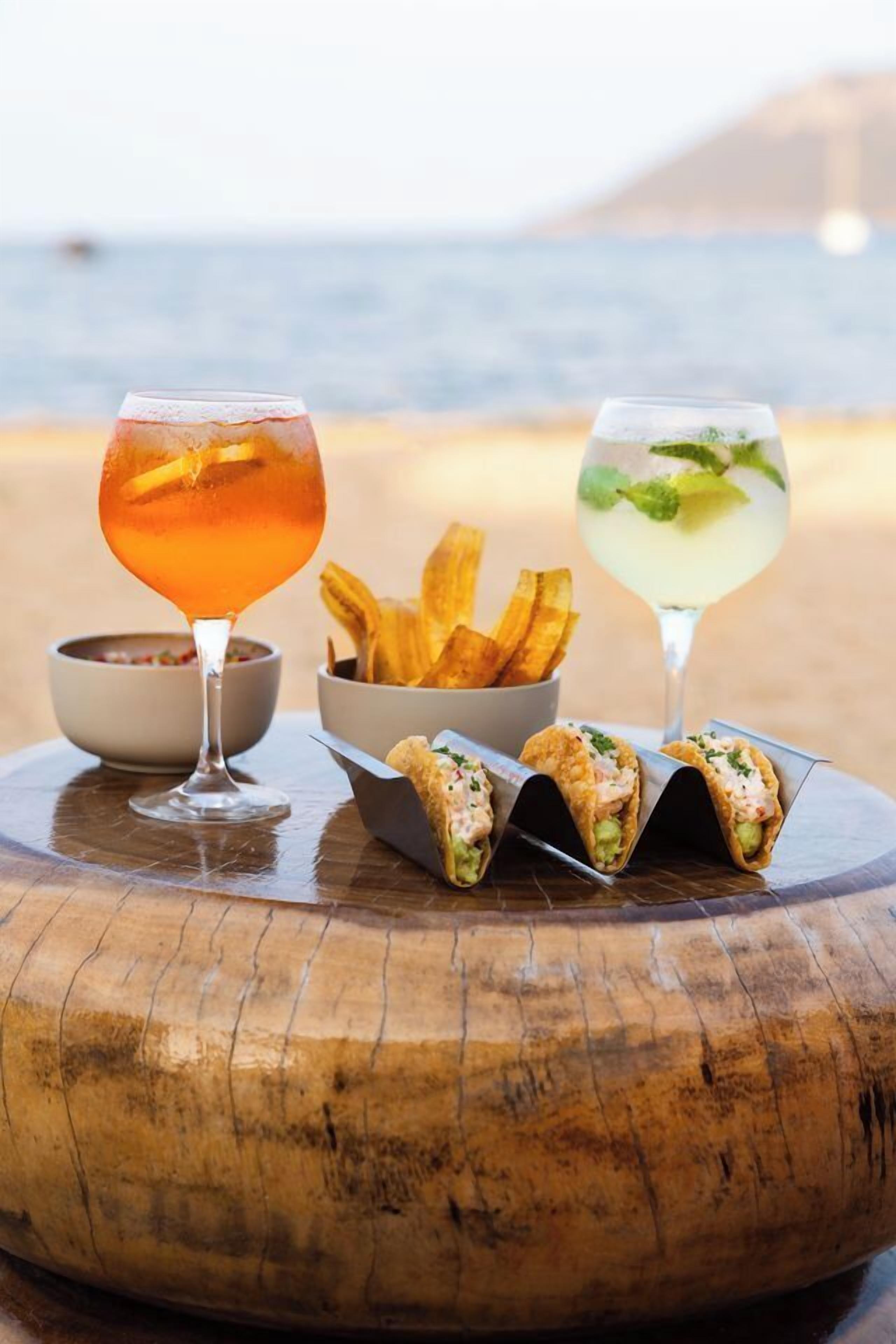 Een houten tafel met een drankje, chips en taco's op het strand.