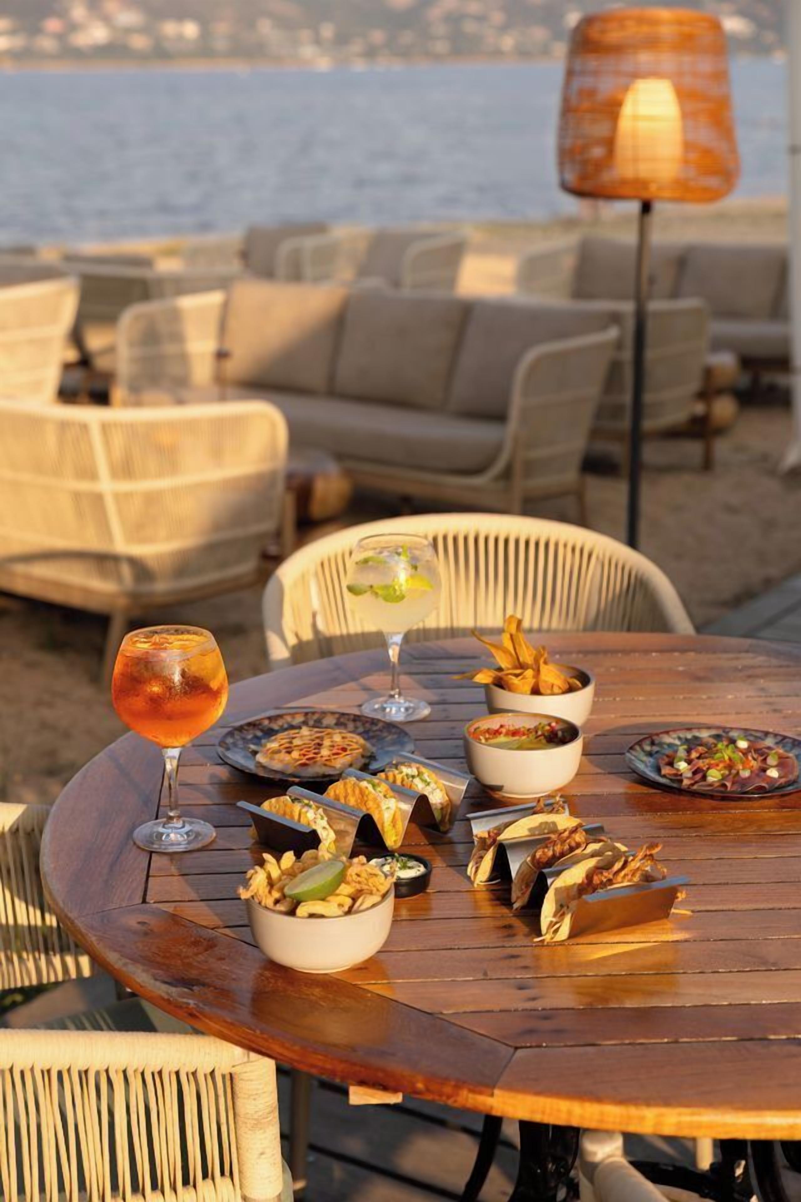 Una mesa redonda de madera con diversos platos y bebidas, rodeada de sillas de mimbre, frente a un pintoresco paisaje marítimo.
