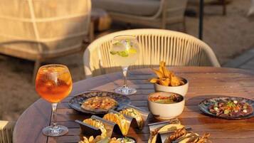 Una mesa redonda de madera con diversos platos y bebidas, rodeada de sillas de mimbre, frente a un pintoresco paisaje marítimo.