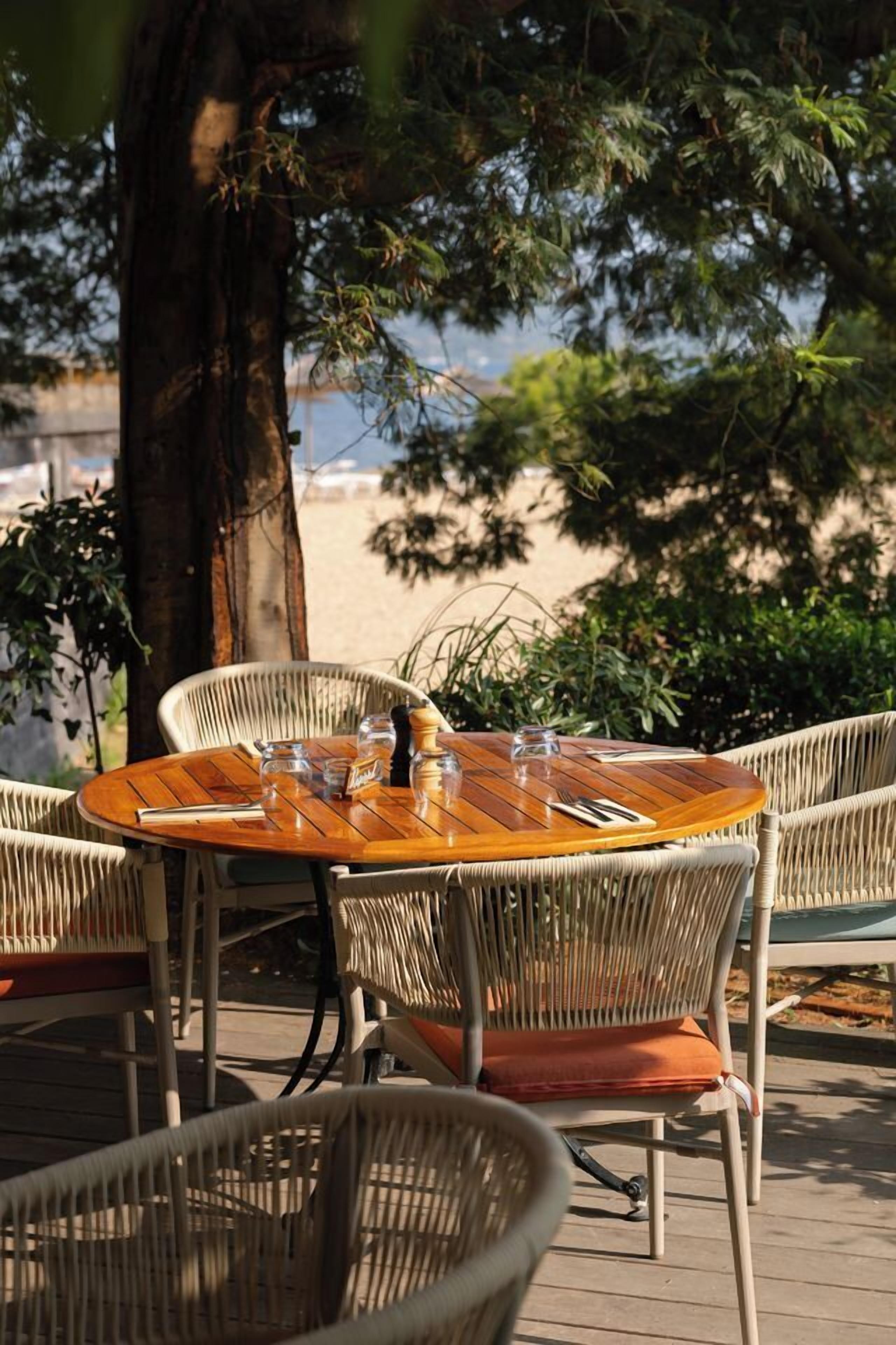 A round wooden table with chairs, set on a wooden deck with a view of a beach and trees in the background.