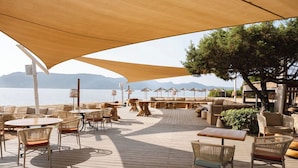 A wooden deck with outdoor seating, including round tables and wicker chairs, overlooking a body of water with mountains in the background. - Hotel Marina Corsica (Porto-Vecchio)