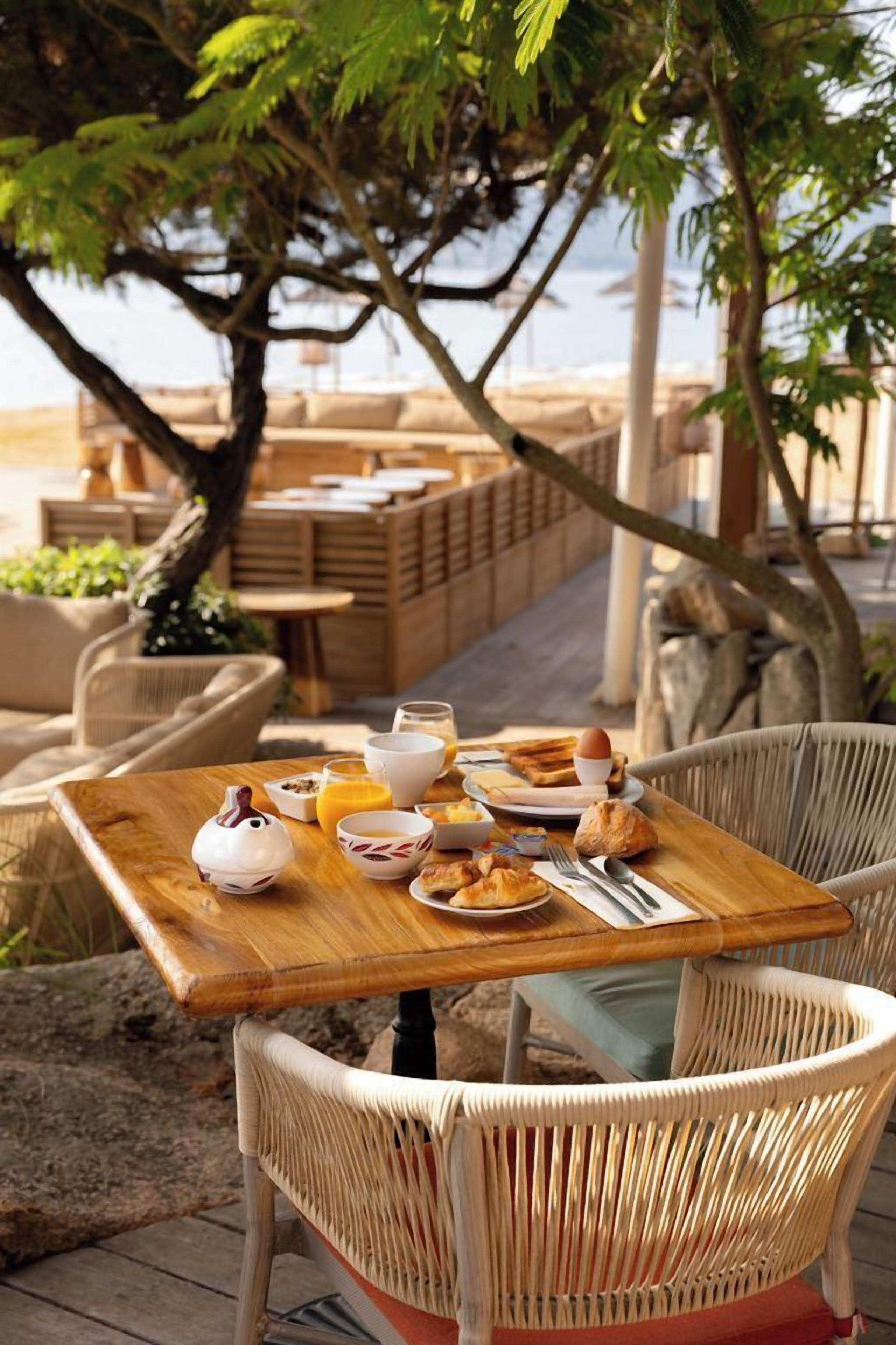 Ein Holztisch mit Frühstücksangeboten, Korbstühle und Blick auf den Strand.