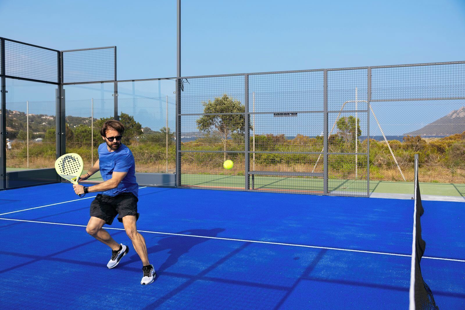 A tennis court with a player in action, a net, and a clear sky in the background.