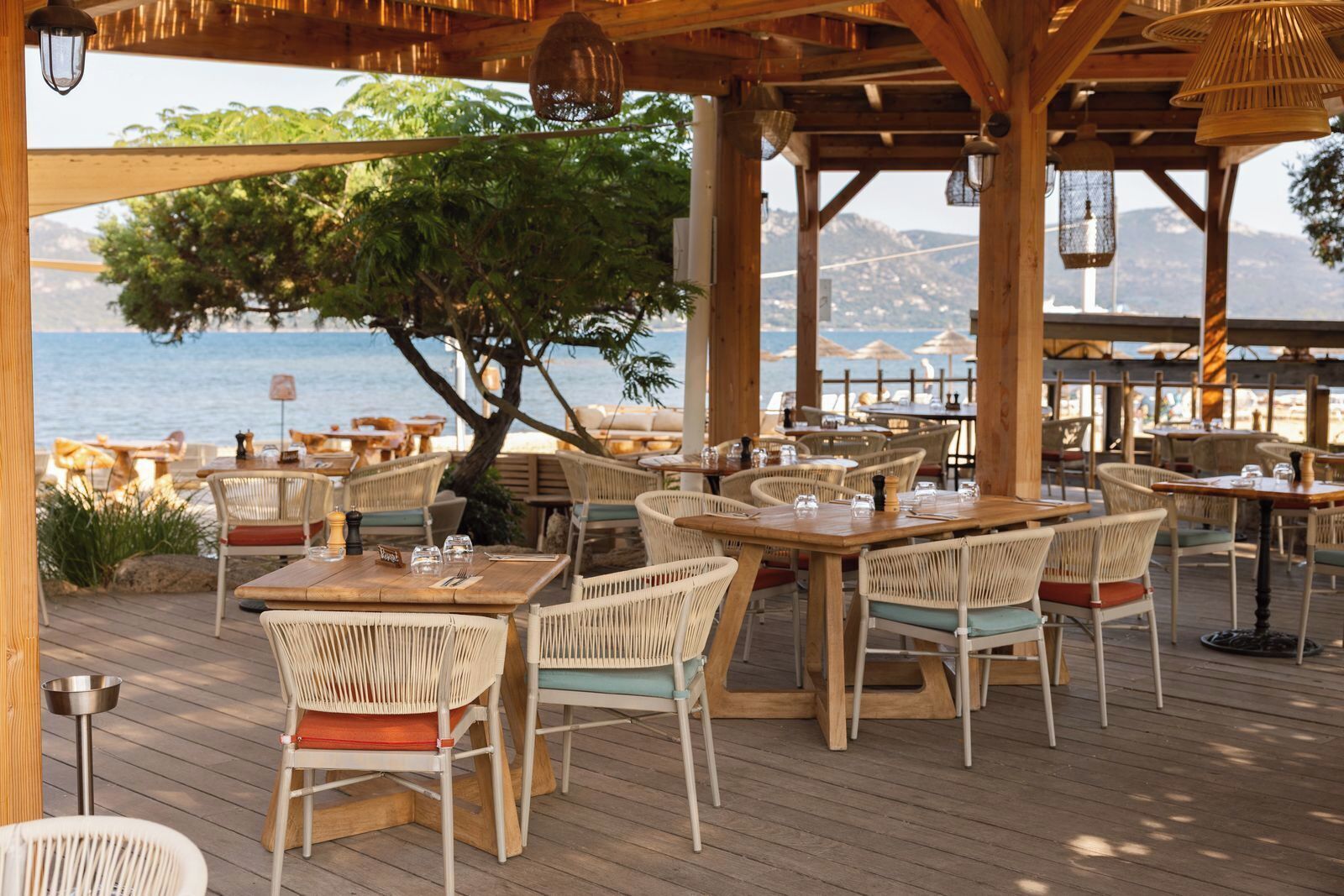 Un espace de restauration en plein air, avec des tables en bois et des chaises en osier, offrant une vue sur un plan d’eau et des montagnes.