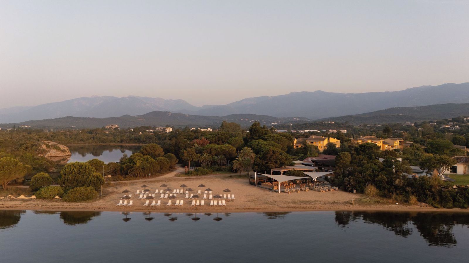 Un resort junto al lago con tumbonas, una zona con sombra y vistas a montañas distantes.
